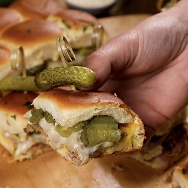 A hand holding a Cuban slider roll with a pickle on top and more sliders below.